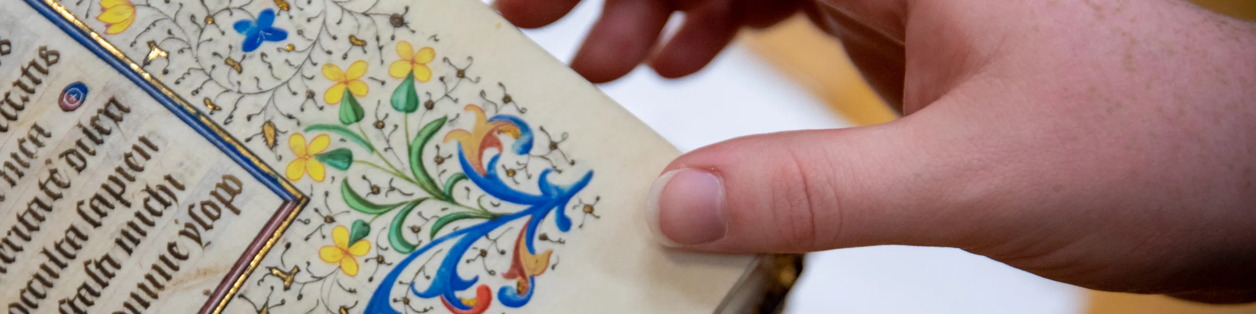 A KU students hand turns the page of an illuminated medieval manuscript with Latin text and gold-leaf floral decoration at the University of Kansas.