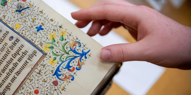 A KU students hand turns the page of an illuminated medieval manuscript with Latin text and gold-leaf floral decoration at the University of Kansas.