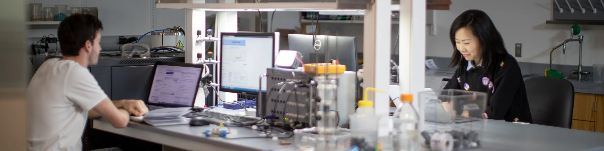 Two KU students work at computer workstations in a research laboratory, reviewing data on laptop and desktop screens.