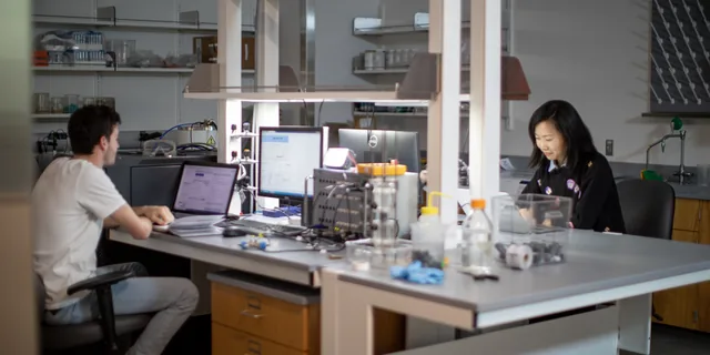 Two KU students work at computer workstations in a research laboratory, reviewing data on laptop and desktop screens.