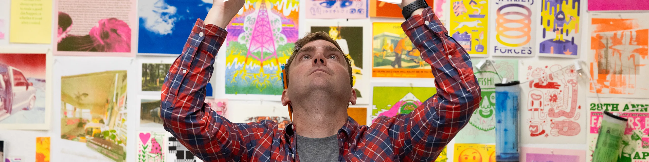 A KU student reviews a printed design in a studio surrounded by colorful posters, illustrations, and graphic artwork, showcasing the creative process and visual communication skills developed in the design degree program.