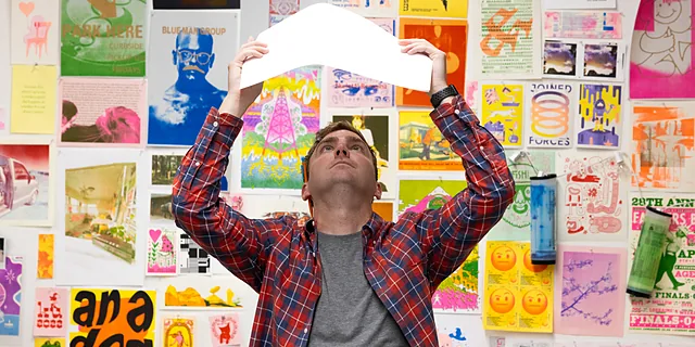 A KU student reviews a printed design in a studio surrounded by colorful posters, illustrations, and graphic artwork, showcasing the creative process and visual communication skills developed in the design degree program.