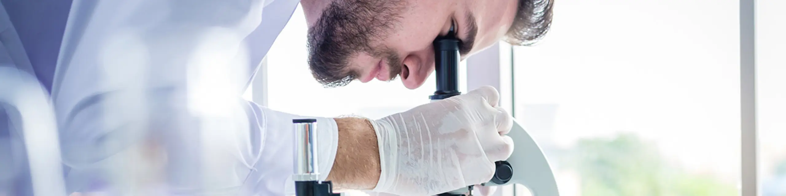 A KU researcher in a white lab coat and gloves looking at a medical sample through a microscope in a laboratory.