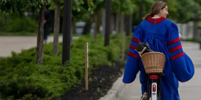 Master of philosophy graduate riding a bike in cap & gown on KU's campus