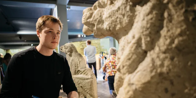 A KU graduate student sketches in a notebook while studying carved Eurasian stone figures displayed in a Slavic museum gallery.